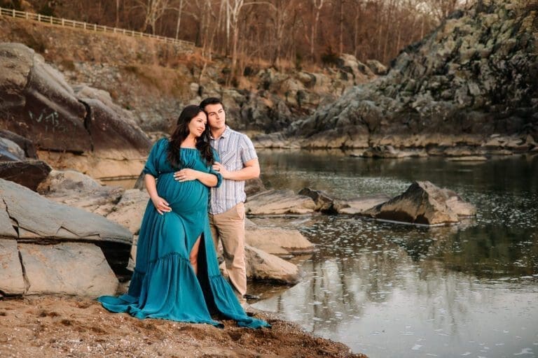 Expectant couple on the edge of the water at Great Falls Park. They lean together and gaze out towards the sunset.