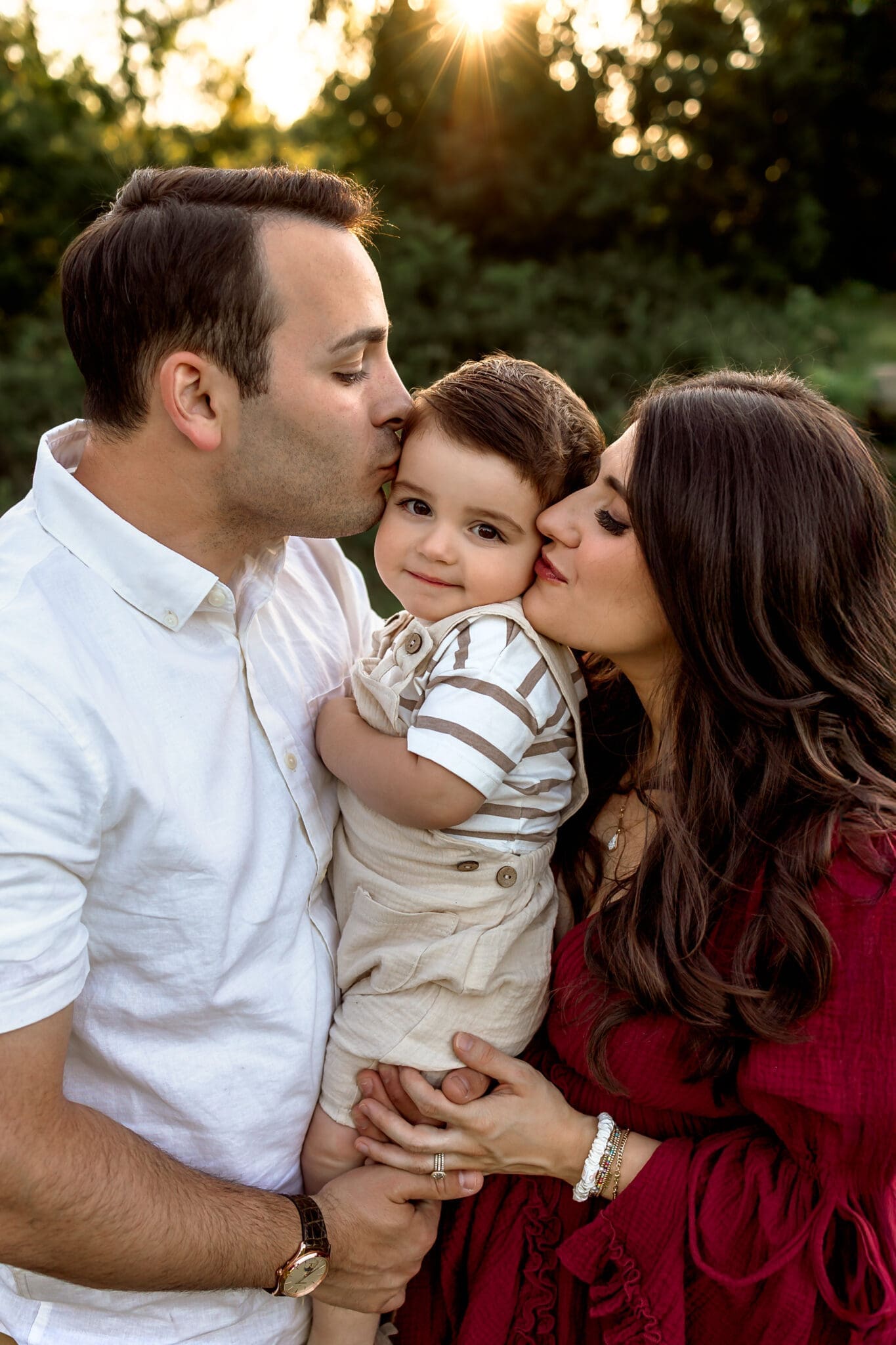 A father in a white shirt kisses his young son’s cheek while the mother, dressed in a red gown, embraces them both in a sunlit field