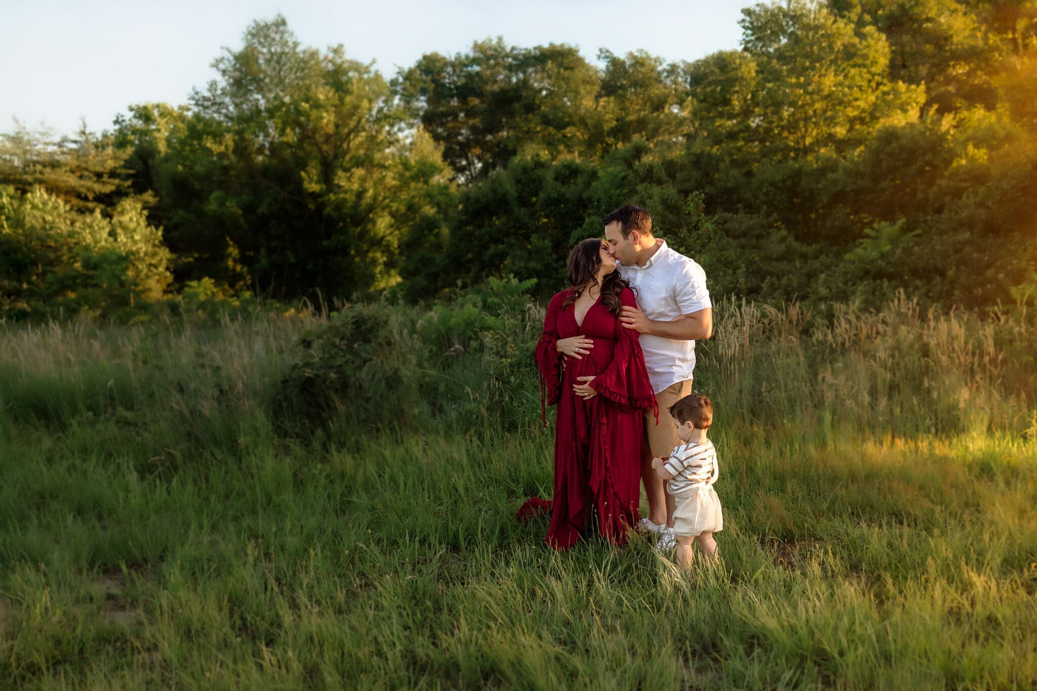 A wide shot of a family of three in a sunlit field, with the father kissing the mother while she holds her pregnant belly and their toddler stands nearby, capturing their spring maternity photos in Annapolis.