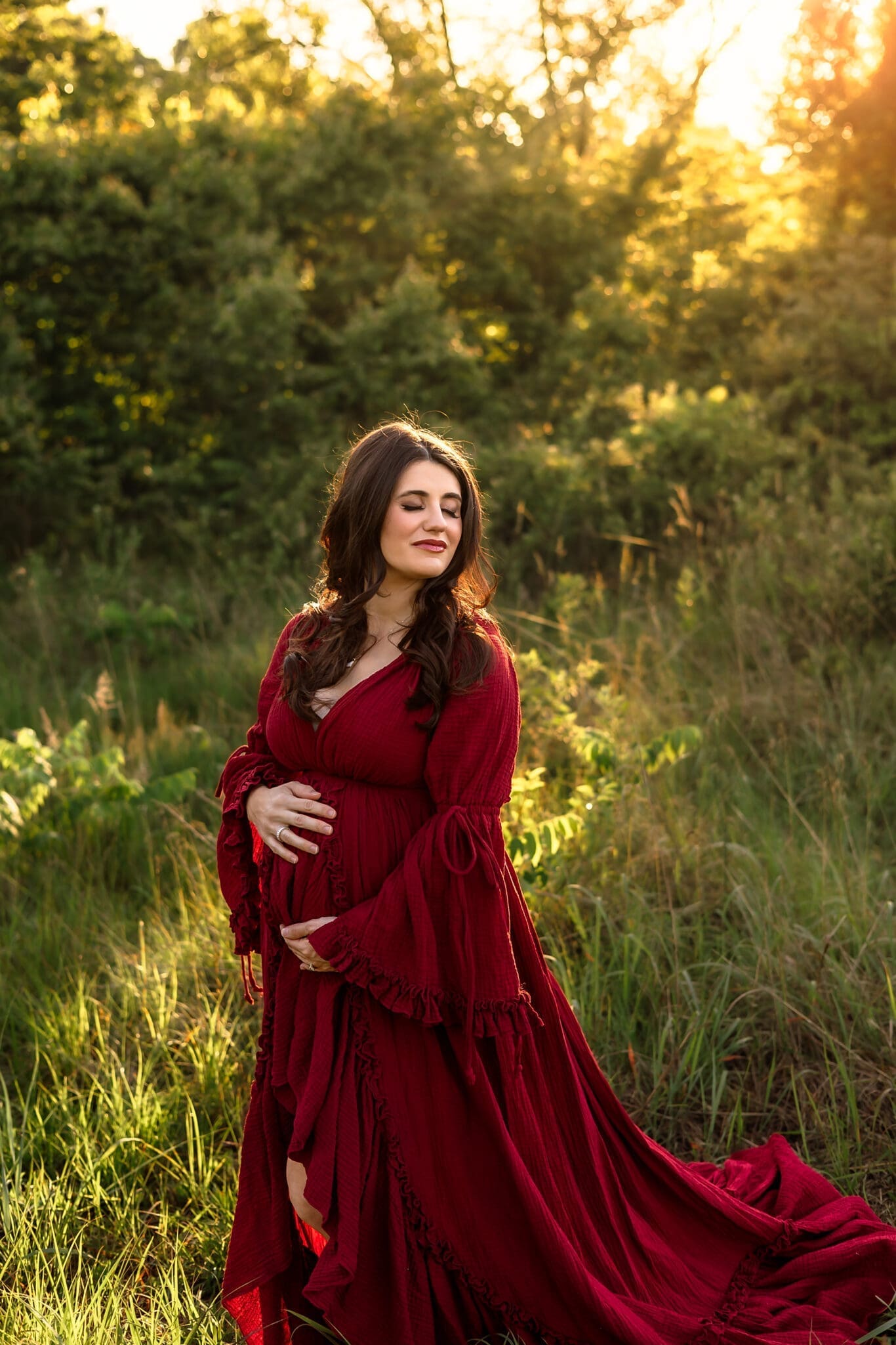 A pregnant woman in a deep red gown stands in a field with her eyes closed, bathed in golden light, during her spring maternity photos in Annapolis.