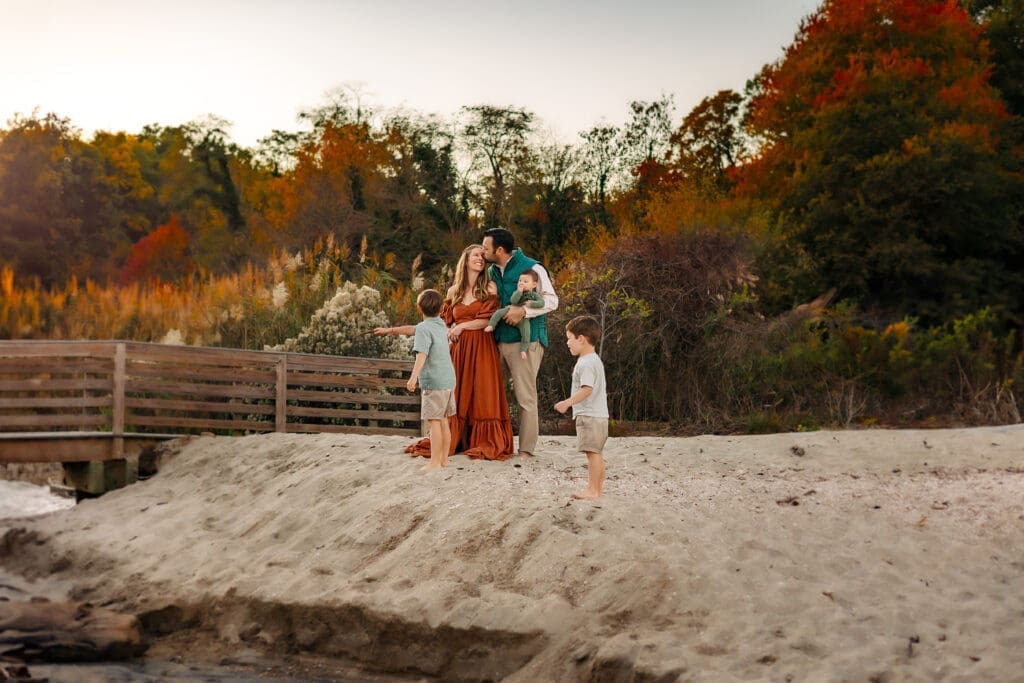 A mom in a rust-colored dress and dad in a teal vest share a kiss on a sandy beach as their two boys look out at the water in this cozy fall mini session.